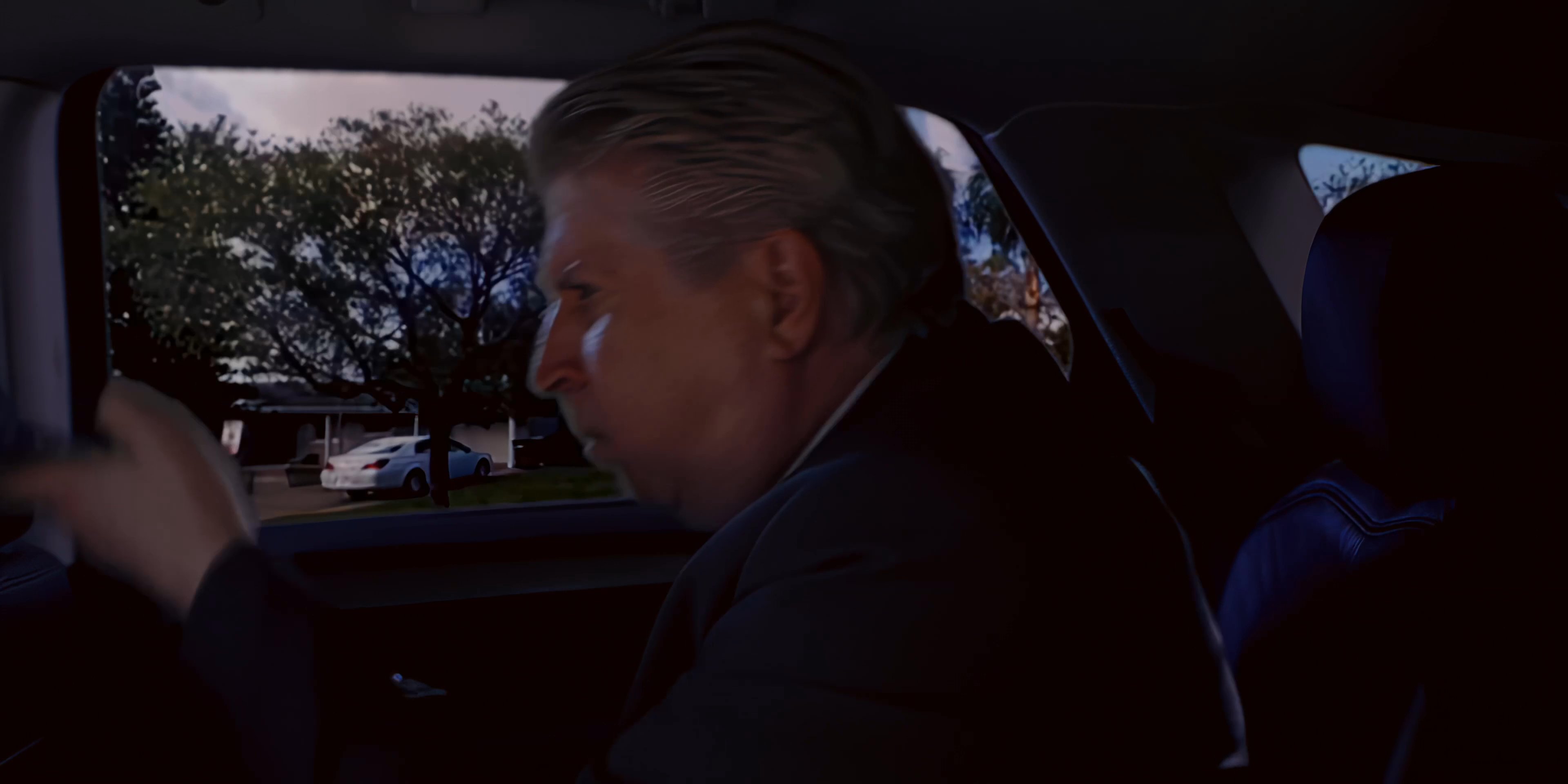 A driver transports a passenger through a suburban neighborhood as the passenger uses a mobile phone in the backseat. The two briefly interact before the driver resumes navigating the road.