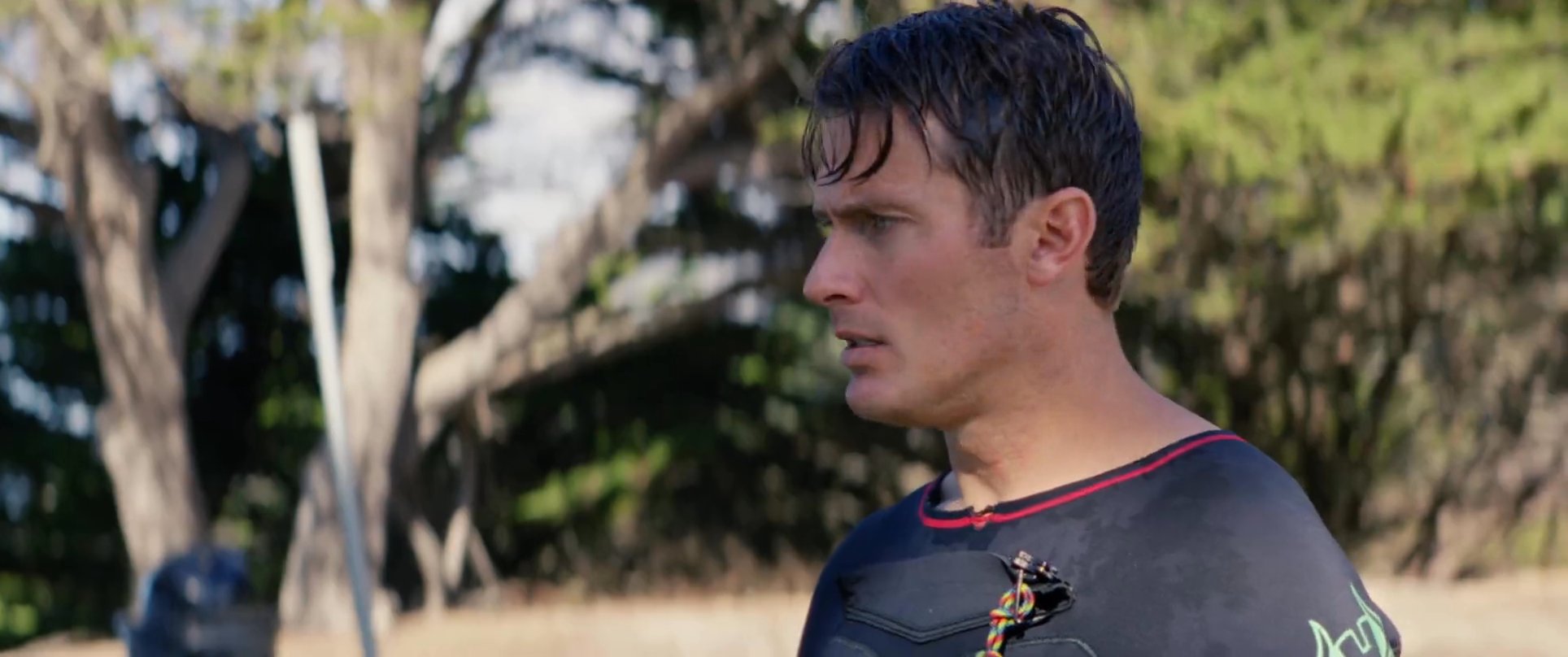 Two individuals in wetsuits stand along a coastal cliff overlooking the ocean, discussing surf conditions before heading to the water.