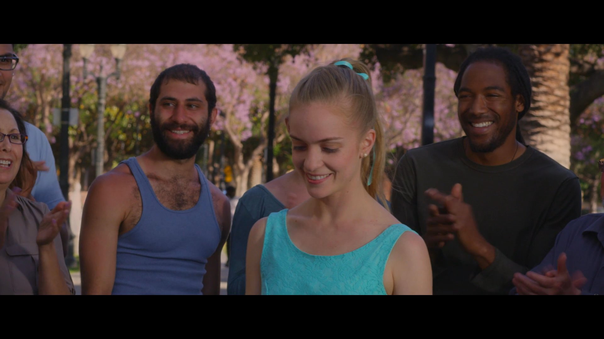 A ballet dancer performs for a small audience in an urban plaza, followed by an ensemble dance sequence. The group interacts positively in a public, tree-lined setting.
