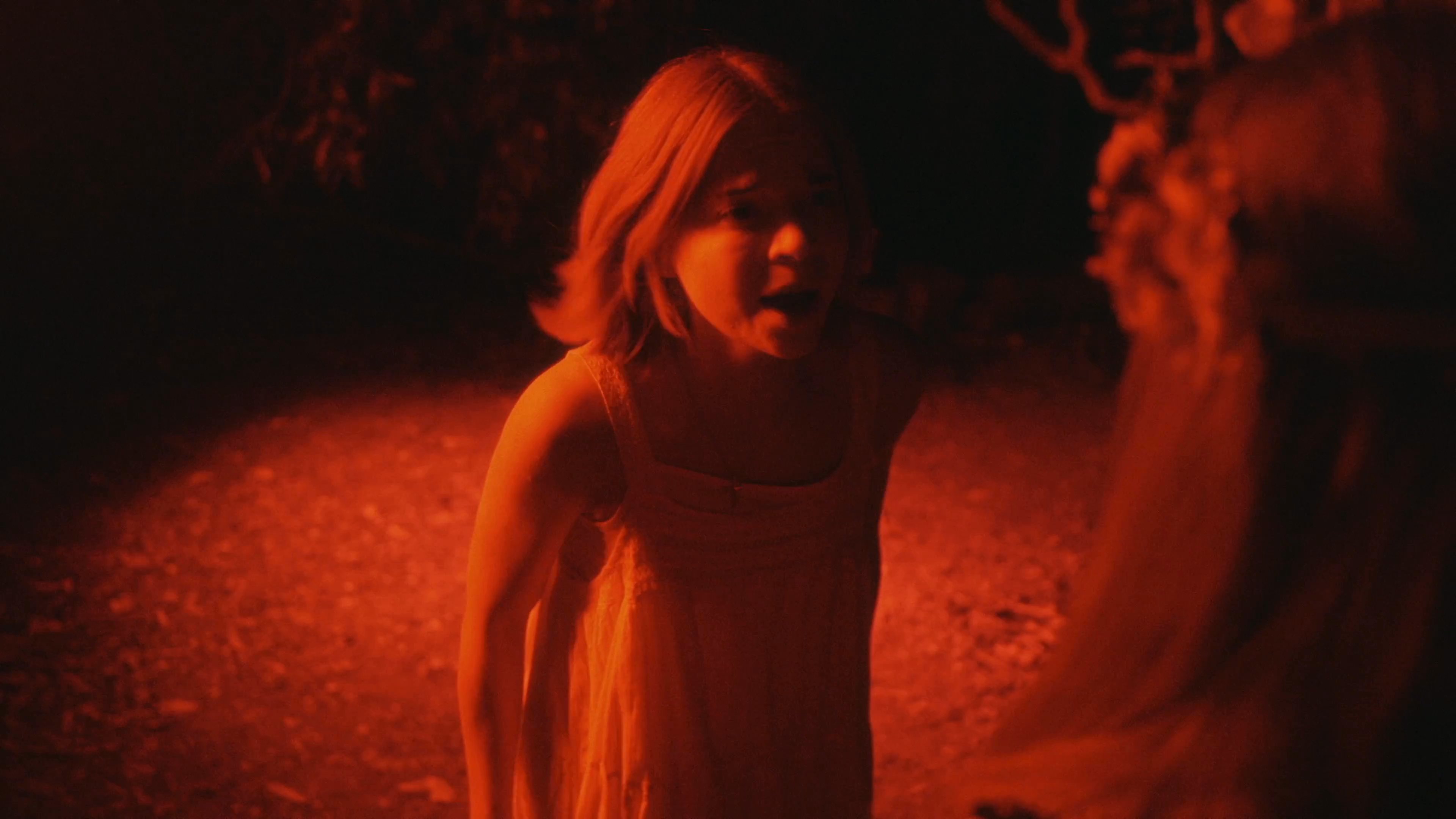 Two individuals interact in a dimly lit, red-hued setting while a blindfolded man remains motionless nearby. The atmosphere suggests a mysterious or ritualistic ceremony unfolding under the cover of darkness.