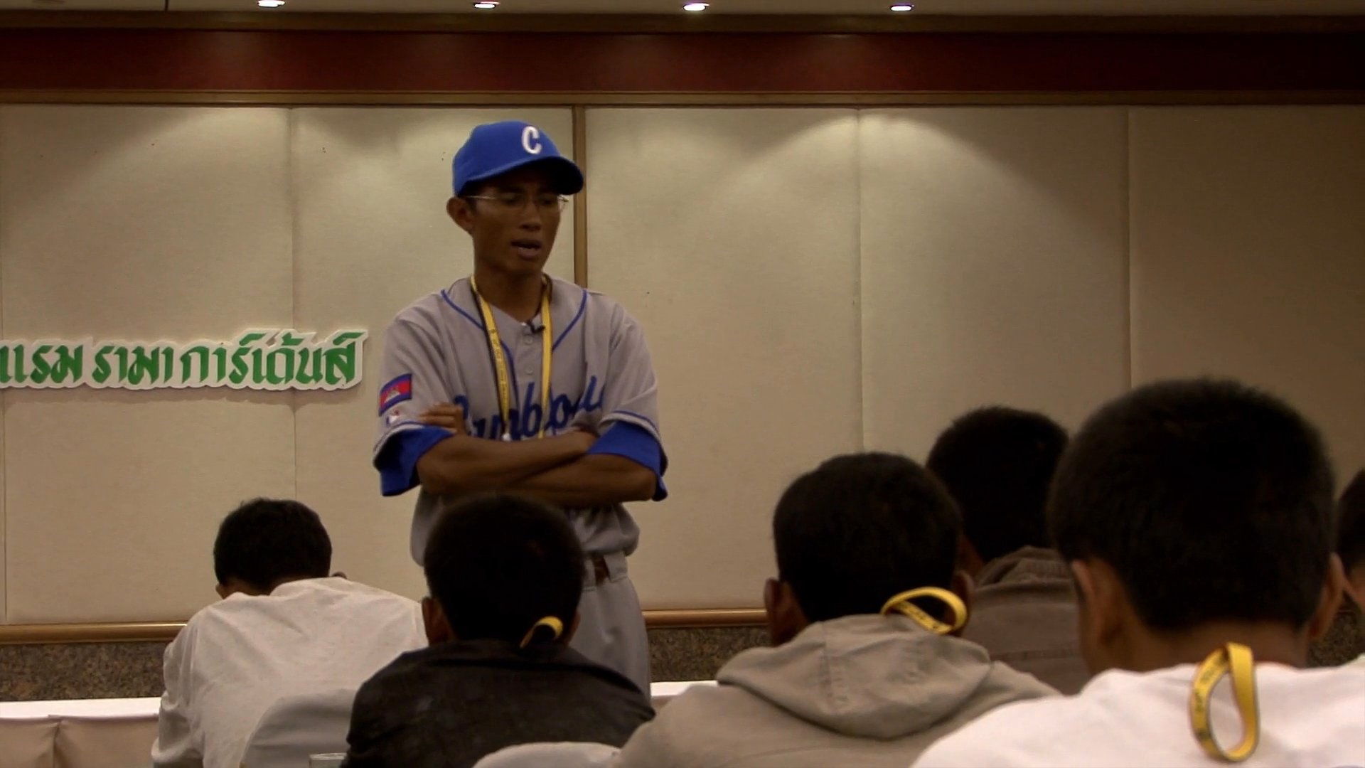 A baseball player in a team uniform delivers a presentation to a group of young athletes in a meeting room, following earlier travel scenes.