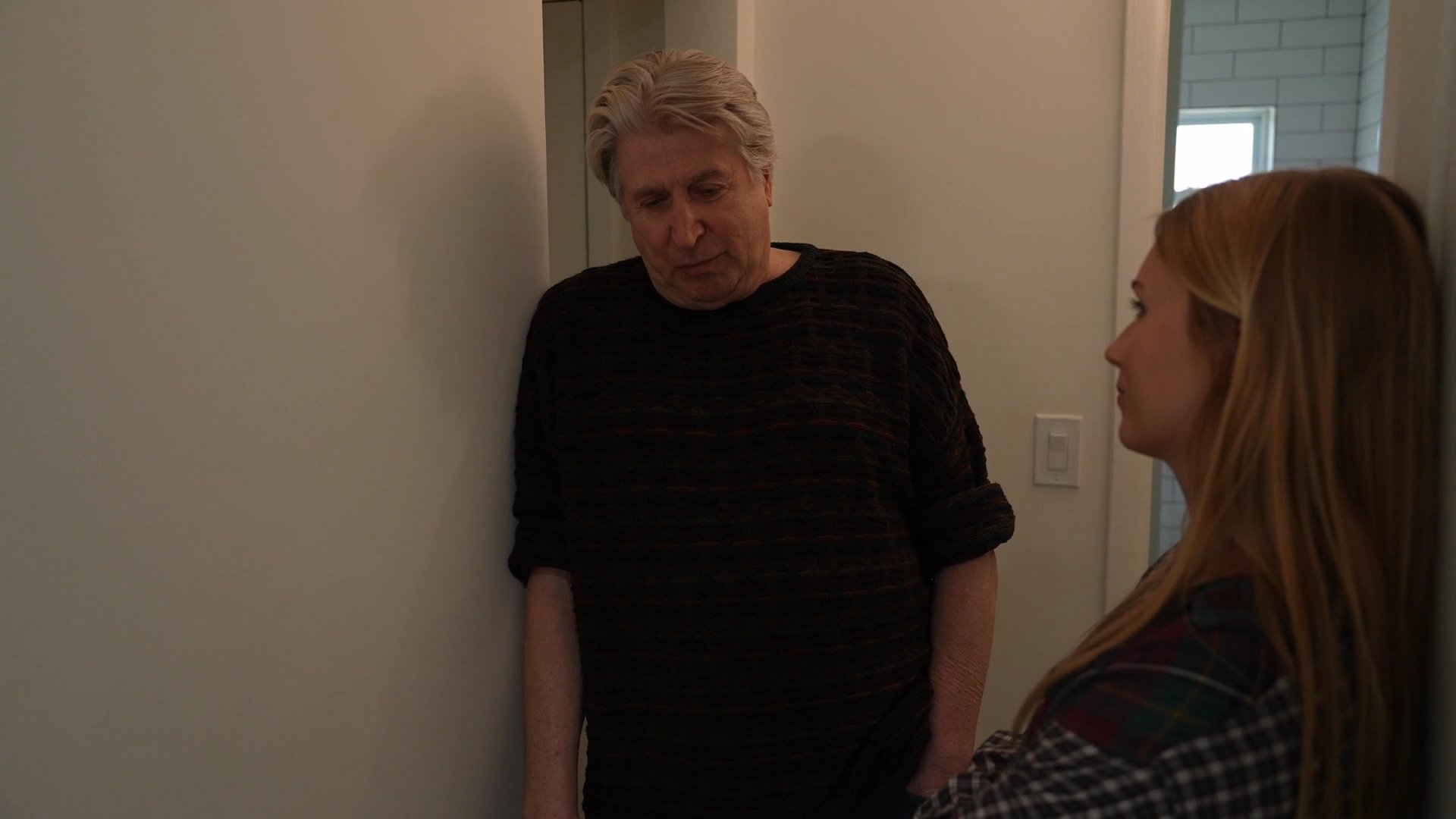 An older man and a young woman engage in a focused, ongoing conversation within a residential corridor.