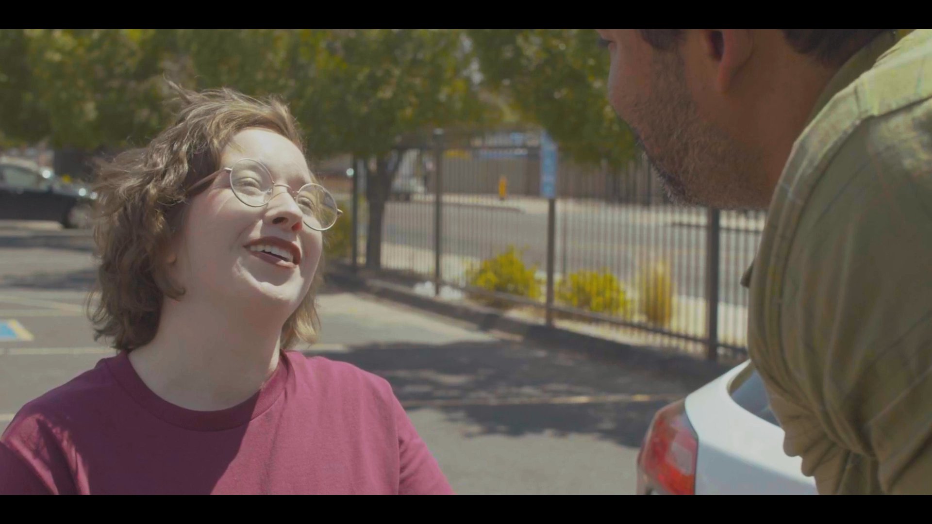 A man using crutches engages in a supportive and intimate conversation with a woman in an outdoor parking lot setting. The interaction emphasizes mutual affection and emotional connection as they communicate.