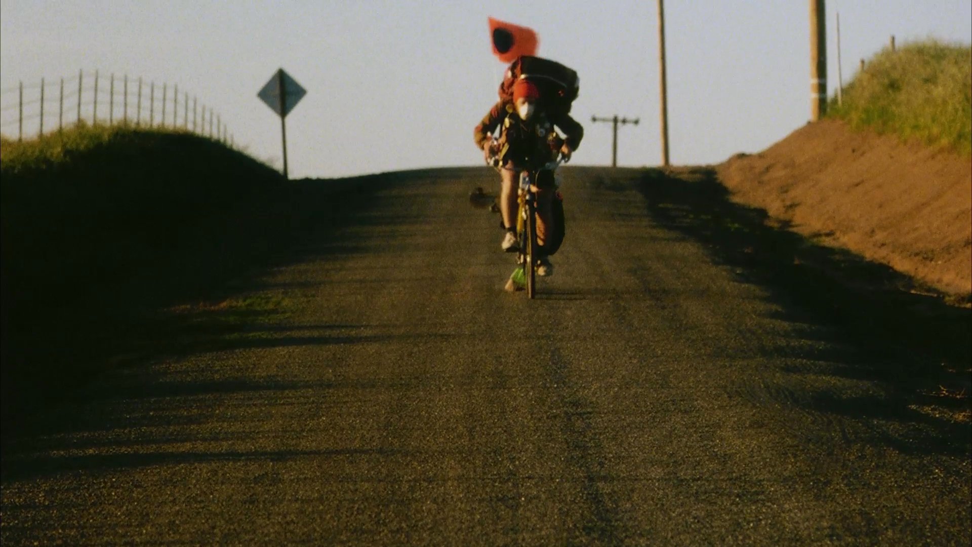 A long-distance cyclist travels down a rural road, passing a large frog resting on the pavement. The journey continues with a frog-themed accessory displayed on the bicycle handlebars, concluding with a title card displaying the text Frog Crossing.