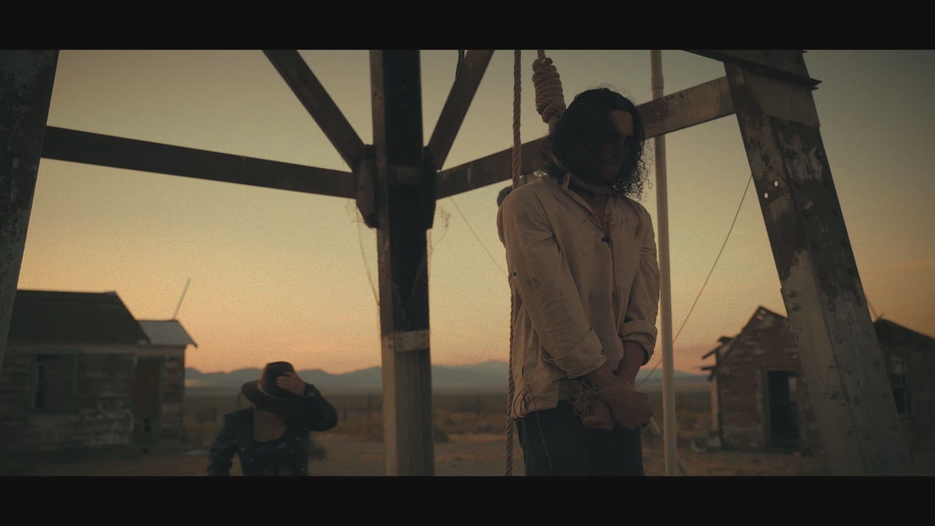 A person hangs beneath a timber gallows in a remote settlement at sunset while another figure crouches nearby. Their tense exchange shifts from an offer to help to a refusal to die, followed by religious exclamations.