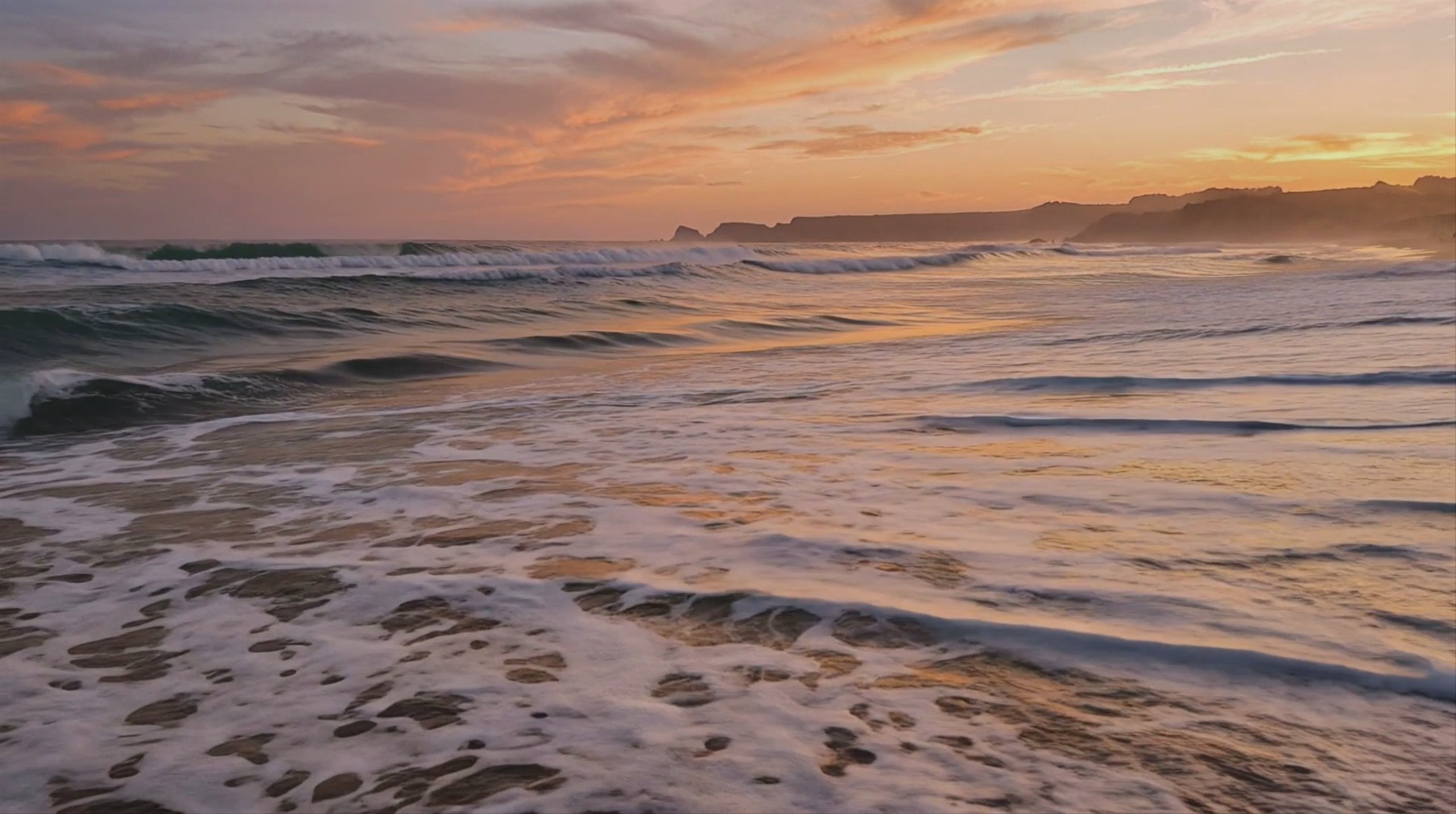 A wide-angle shot of ocean waves crashing onto a sandy beach at sunset, with distant cliffs on the horizon. The orange and pink sky is reflected on the wet shoreline. This 1924x1076 video frame was generated using the Kling 2.1 Master model.