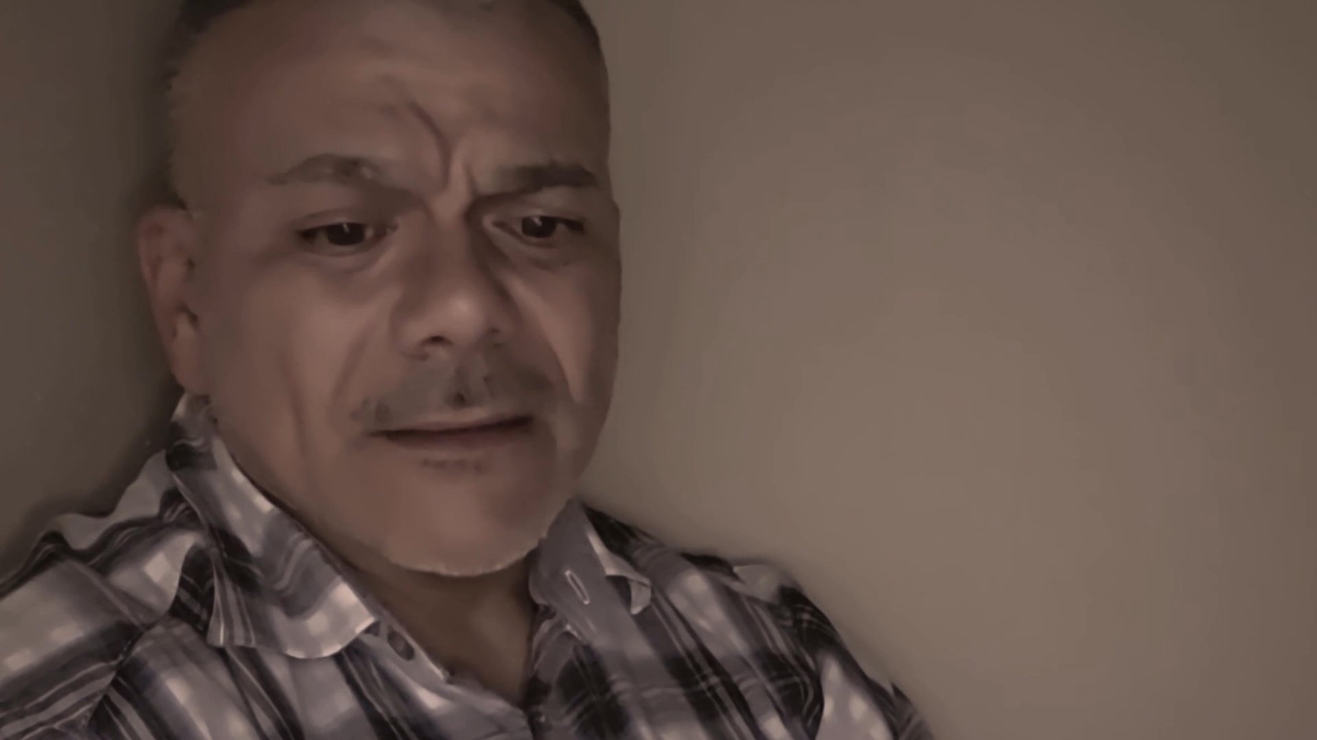 A man wearing a plaid shirt speaks directly to the camera with expressive facial gestures and emotive delivery.