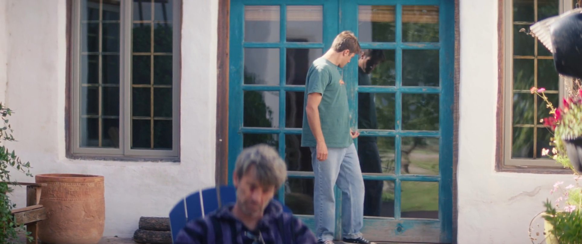 A woman speaks indoors while a man reads a book on a patio nearby. The man subsequently steps out of the house to observe the reader, establishing an atmosphere of quiet reflection.