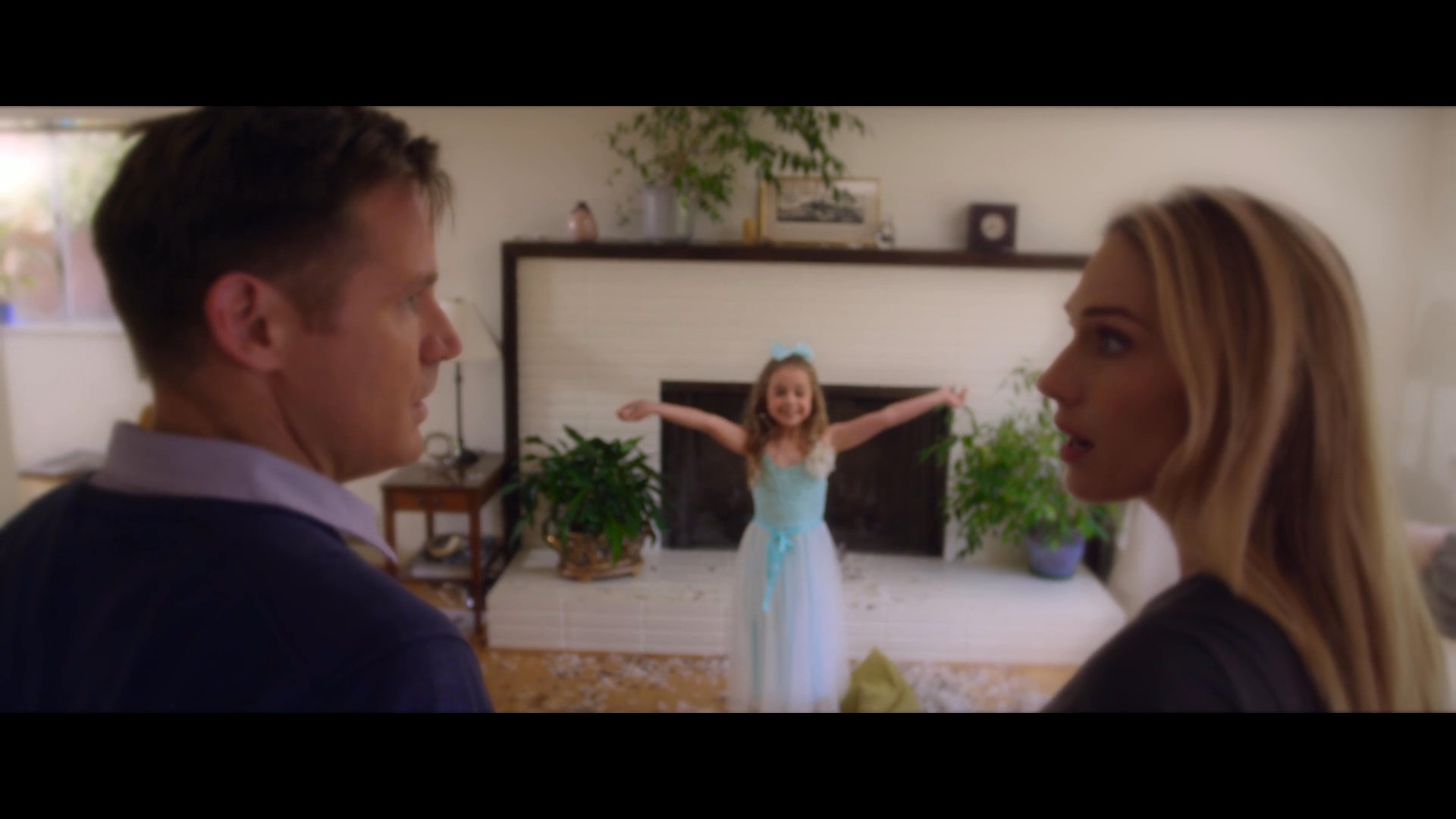 A couple enters a home filled with scattered flower petals before observing their young daughter perform in a ballet studio.