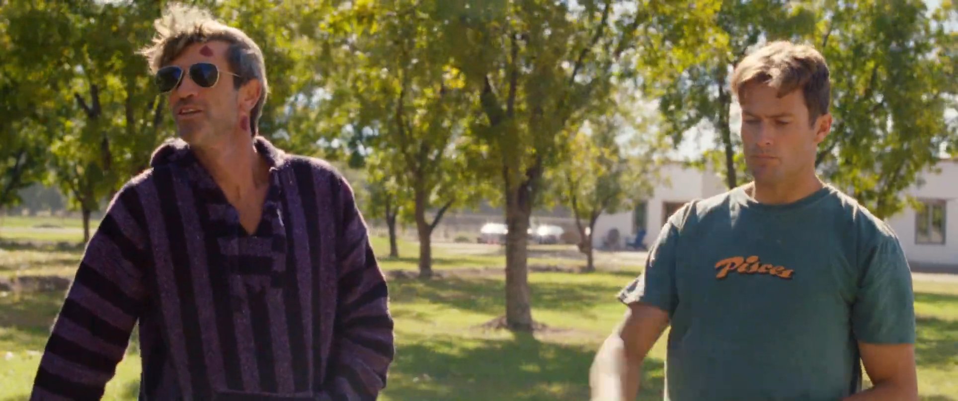 Two men engage in a conversation while standing in a park-like outdoor setting with trees in the background. One man wears a dark striped hoodie and sunglasses, while the other is dressed in a green t-shirt.